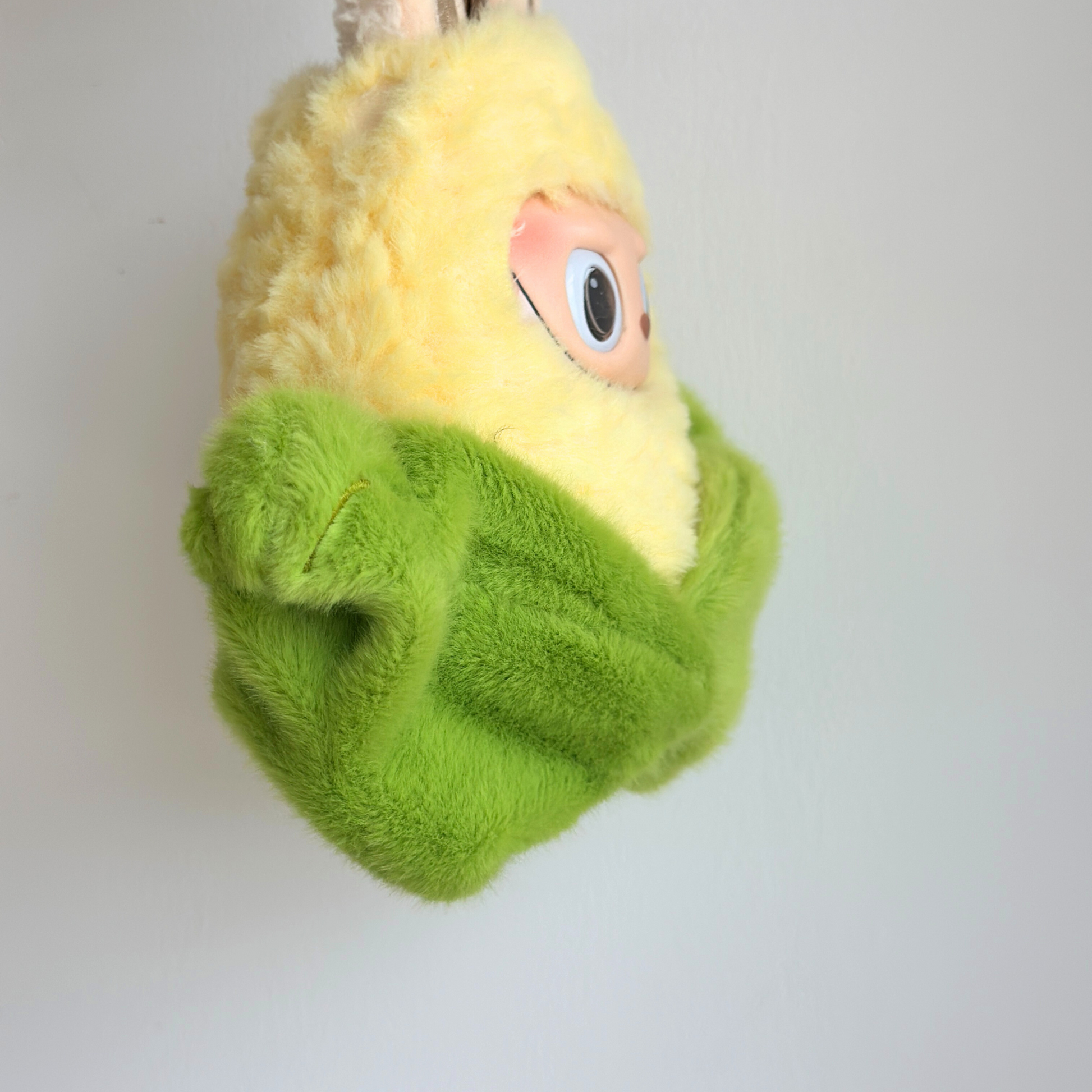 Side angle view of Labubu doll in corn costume showing yellow fuzzy corn kernels hood and green plush corn husk wrapping - detailed vegetable-themed doll outfit from profile perspective