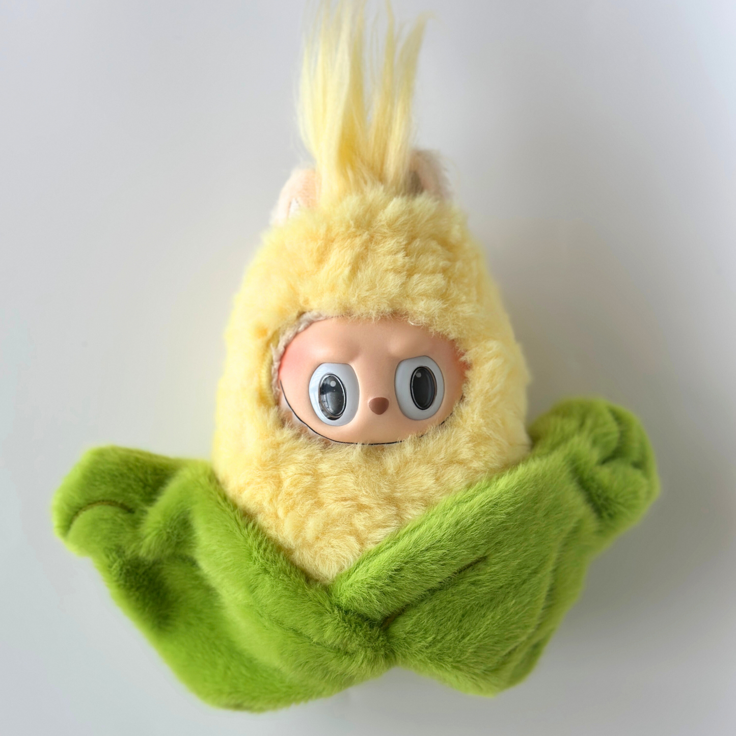 Labubu doll wearing corn costume with yellow fuzzy head representing corn kernels and green plush corn husk leaves wrapped around body - cute vegetable-themed doll outfit