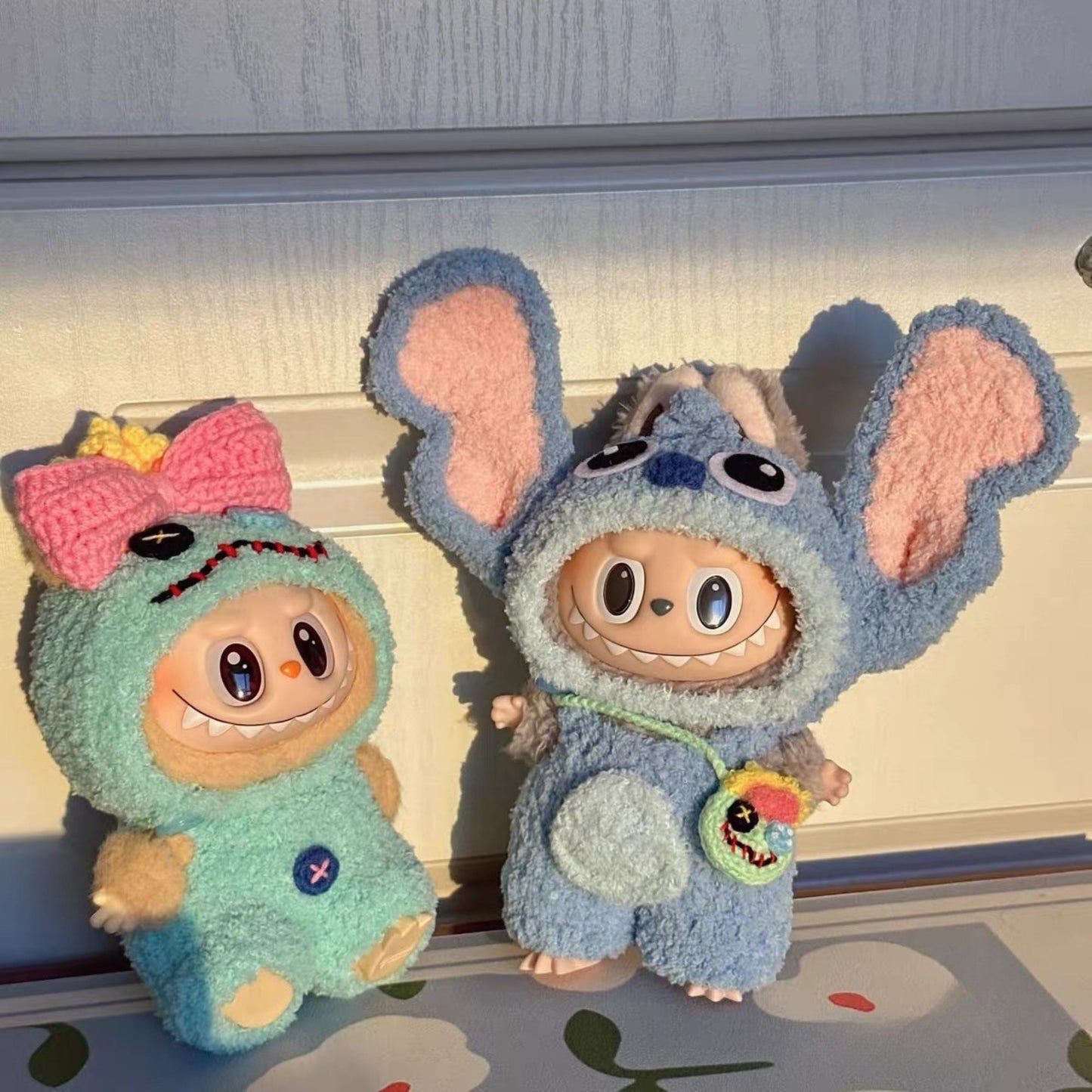 Two labubu dolls one in a green outfit with a pink bow and the other in a blue stitch outfit, on a light-colored surface.