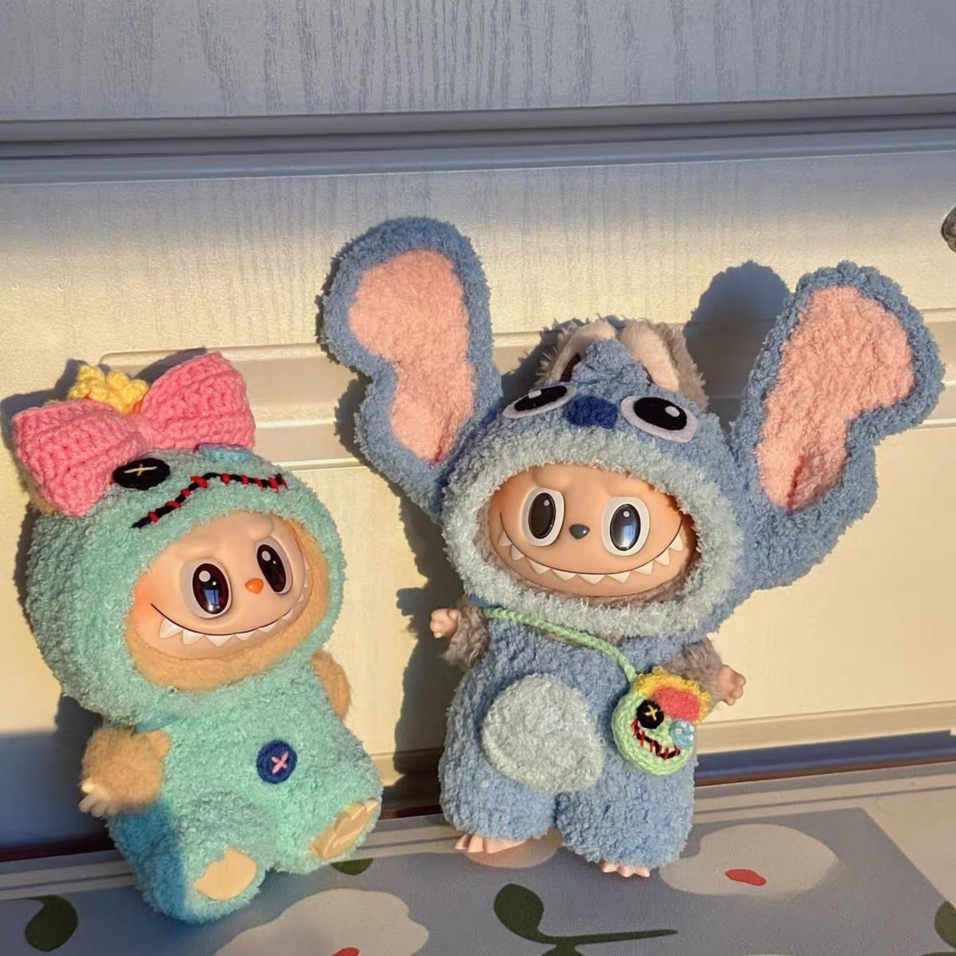 Two labubu dolls one in a green outfit with a pink bow and the other in a blue stitch outfit, on a light-colored surface.