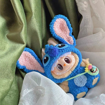 Labubu wearing blue stitch outfit with pink ears, sitting on a textured surface.