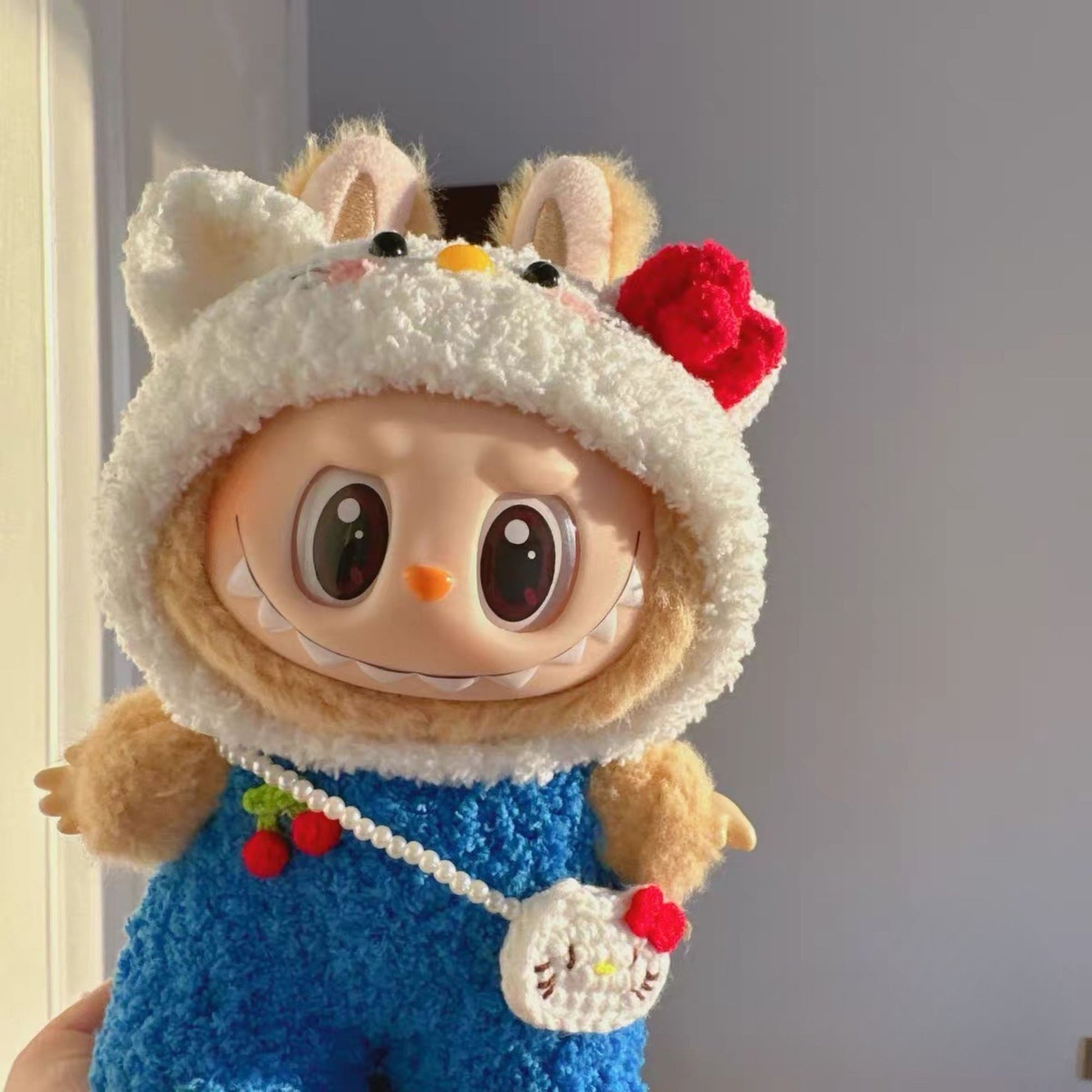 Labubu doll wearing adorable Hello Kitty costume with white fluffy hood featuring cat ears, orange nose, black eyes, red bow, and blue sequined dress with cherry decorations