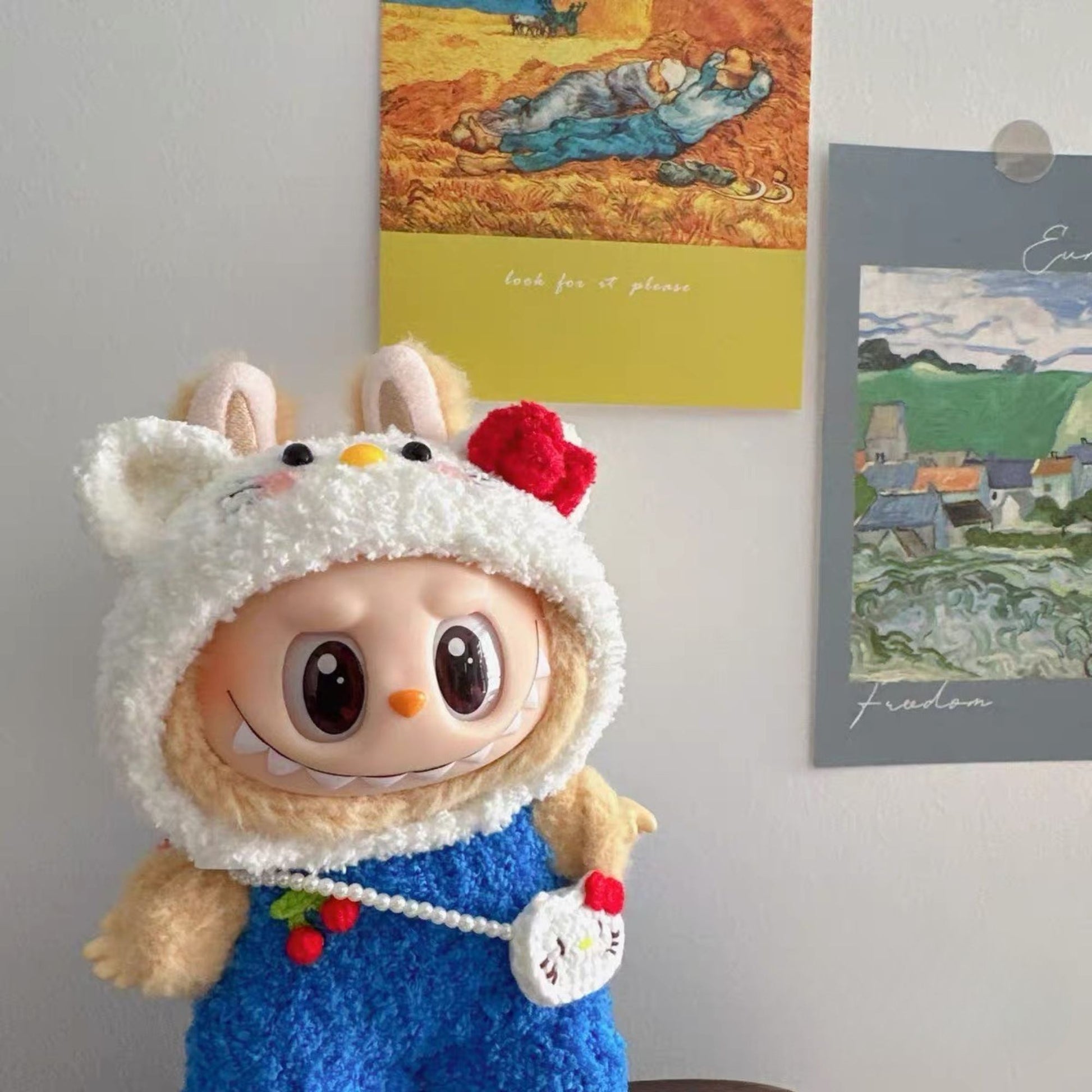 Labubu doll in Hello Kitty costume displayed in cozy room setting with Van Gogh artwork and landscape prints on the wall, showcasing the adorable white cat hood and blue sequined dress