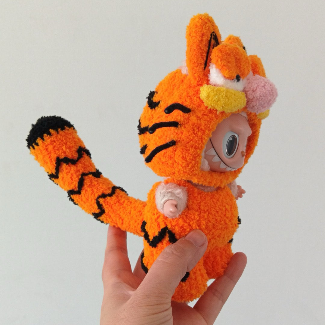 Hand holding a labubu in an orange garfield costume against a plain background