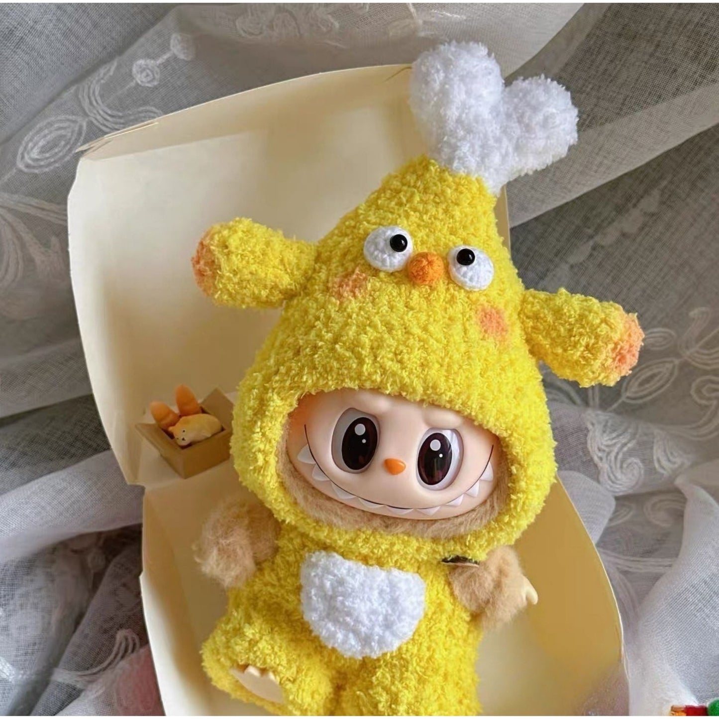 Labubu doll wearing yellow chicken drumstick costume with white fluffy wings, orange accents, and googly eyes, sitting in a cream-colored bowl on decorative lace fabric
