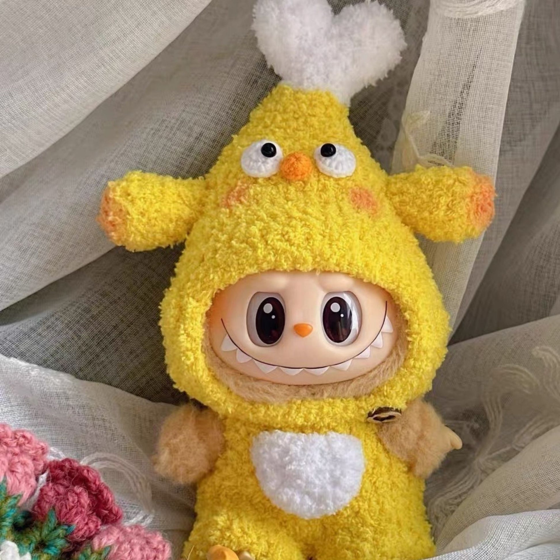 Handcrafted yellow chicken drumstick outfit for Labubu doll featuring fluffy white wing details, orange textured accents, black safety eyes and white belly patch, photographed on delicate white lace with handmade pink crocheted flower decorations