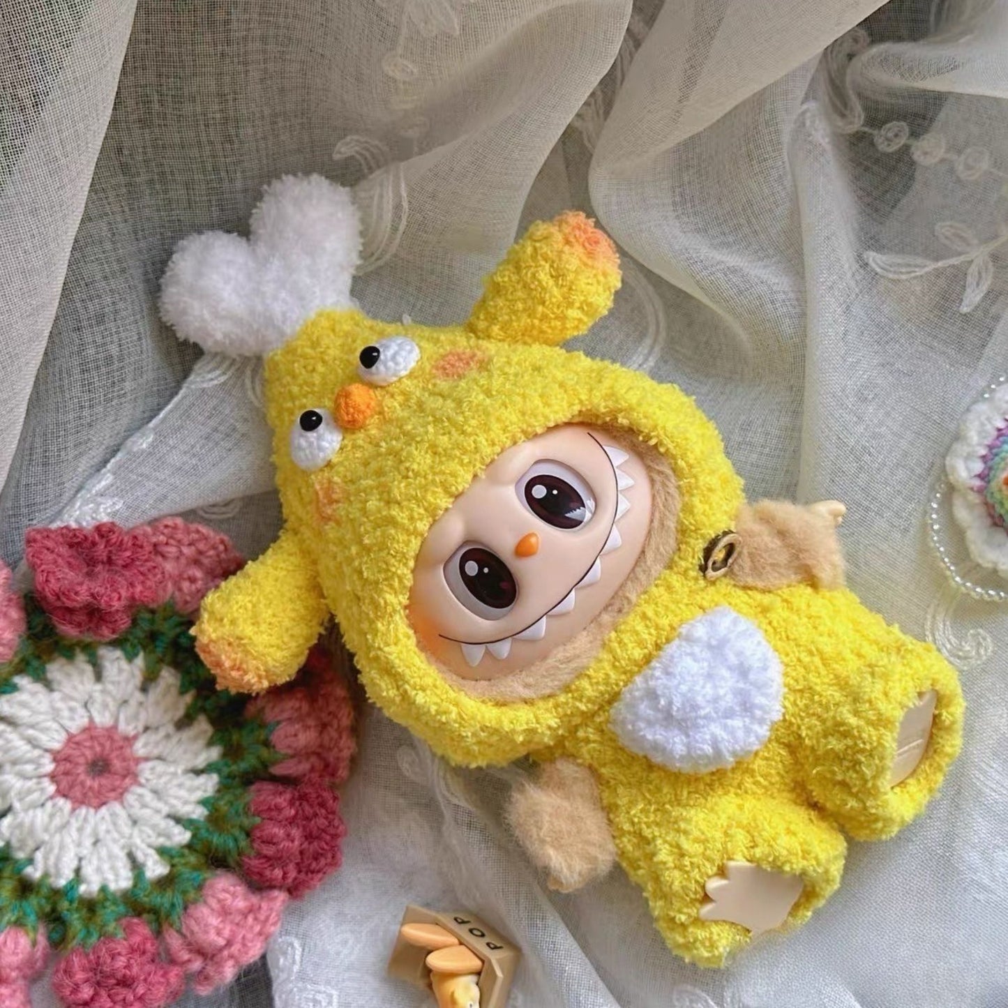 Labubu doll wearing a yellow chicken drumstick costume with white fluffy details, orange beak, and black eyes, displayed on white lace fabric alongside crocheted flowers