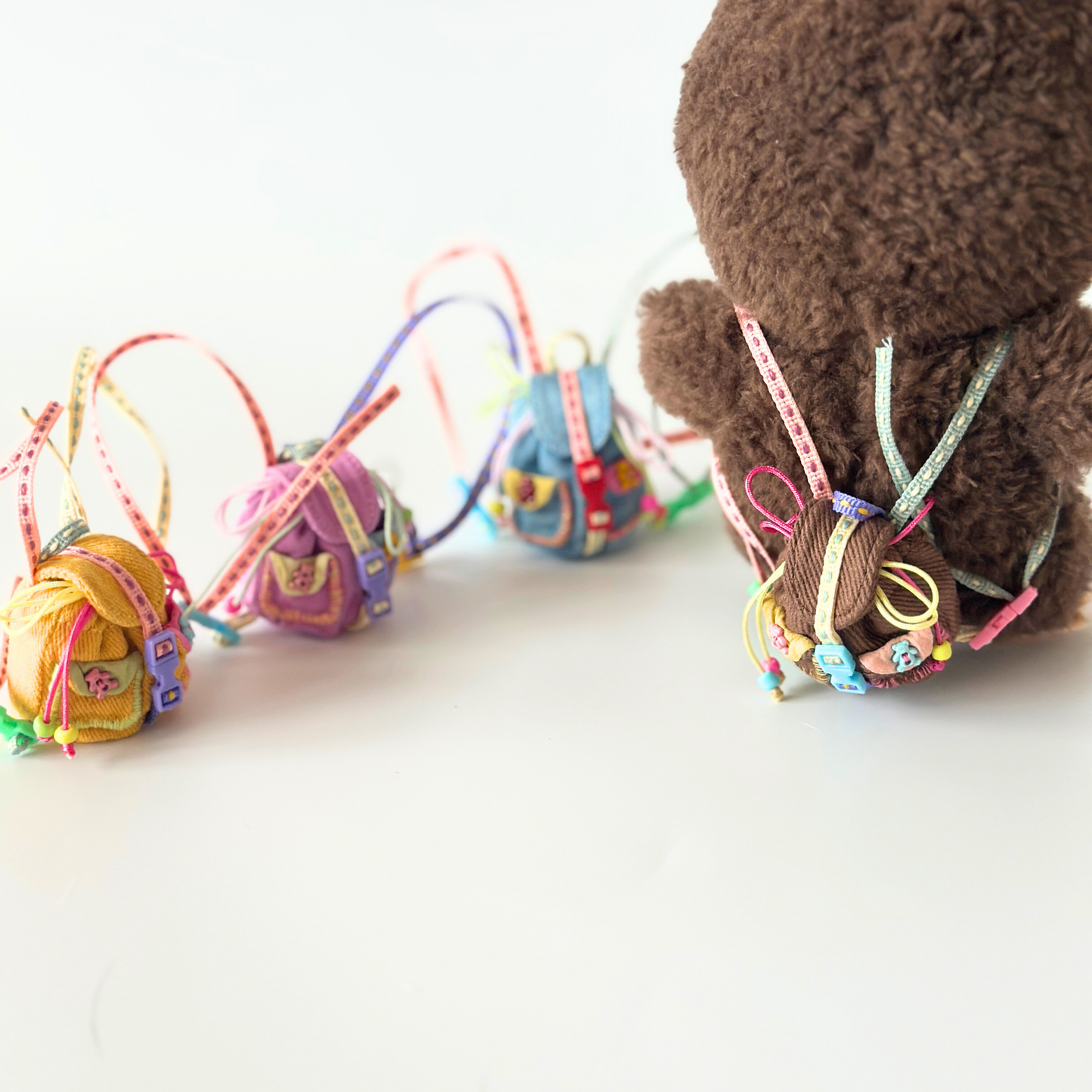 Lifestyle shot of Labubu miniature backpack collection featuring brown Labubu doll wearing corduroy backpack with colorful rainbow straps, alongside other handmade backpacks in yellow, purple, and blue colors. Shows various miniature accessories with metal buckles, decorative ribbons and charms scattered in artistic arrangement, demonstrating quality craftsmanship and variety for collectors.