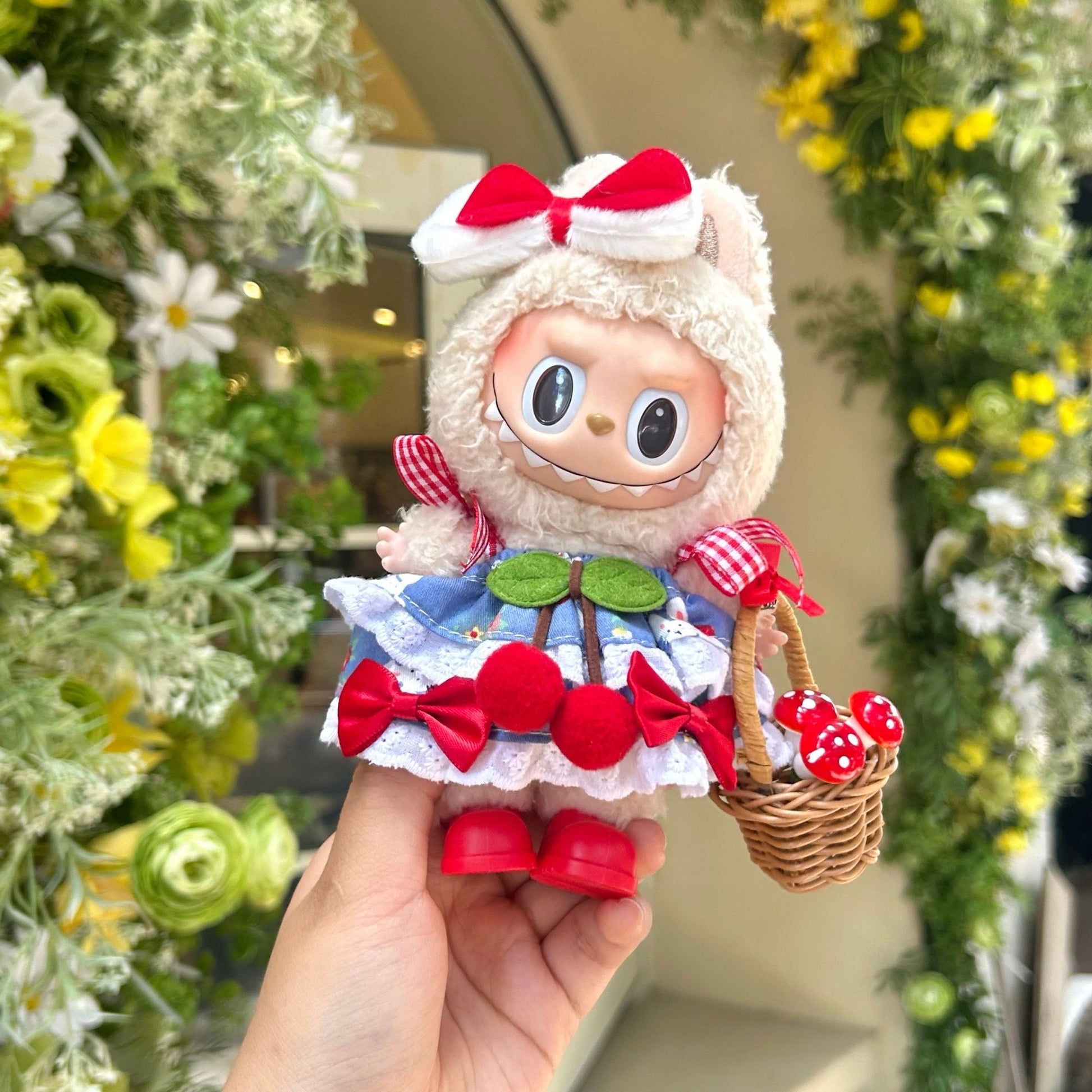 Hand holding Labubu doll wearing handmade cherry-themed costume featuring red bow headband, blue dress with white lace collar, red gingham ribbon sleeves, cherry decorations and red bow accents on skirt, holding miniature wicker basket filled with red mushroom accessories. Set against natural flower garden background, perfect themed doll outfit for collectors and craft enthusiasts.