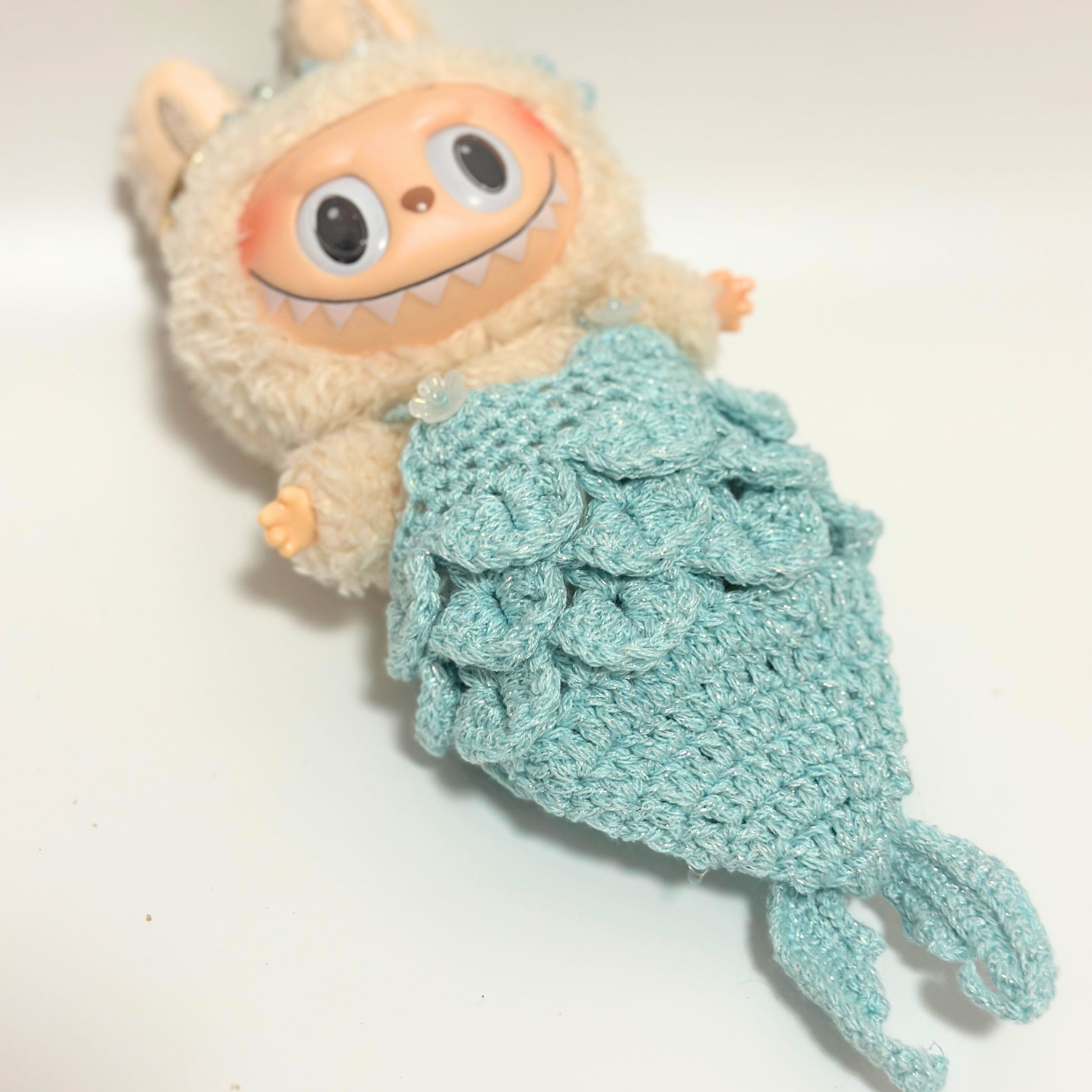 Side angle view of Labubu doll wearing handmade crocheted mermaid costume in mint green showcasing detailed dimensional scale texture, layered scale pattern creating realistic mermaid tail silhouette. Features intricate crochet craftsmanship with overlapping scales and tapered tail design, perfect fantasy-themed collectible doll costume for mermaid enthusiasts and toy collectors.
