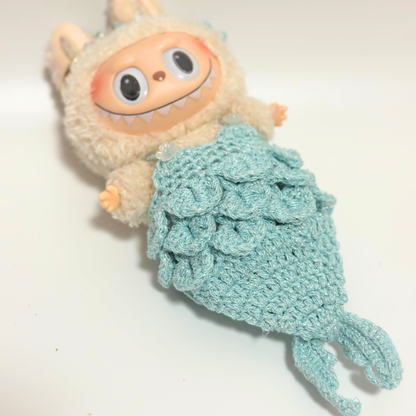 Side angle view of Labubu doll wearing handmade crocheted mermaid costume in mint green showcasing detailed dimensional scale texture, layered scale pattern creating realistic mermaid tail silhouette. Features intricate crochet craftsmanship with overlapping scales and tapered tail design, perfect fantasy-themed collectible doll costume for mermaid enthusiasts and toy collectors.