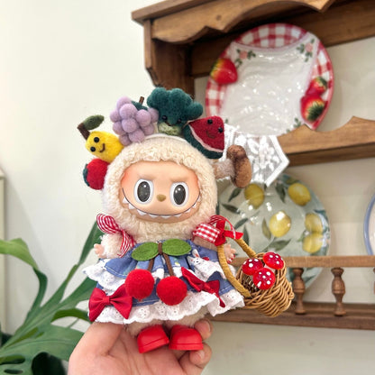 Hand holding Labubu doll wearing handmade fruit-themed costume featuring colorful fruit crown with grapes, broccoli, watermelon, lemon and strawberry decorations, blue dress with white lace collar, red gingham ribbon sleeves, cherry decorations and red bow accents on skirt, holding miniature wicker basket with red polka dot mushrooms. Set against cozy kitchen background with decorative plates, perfect themed doll outfit for collectors.
