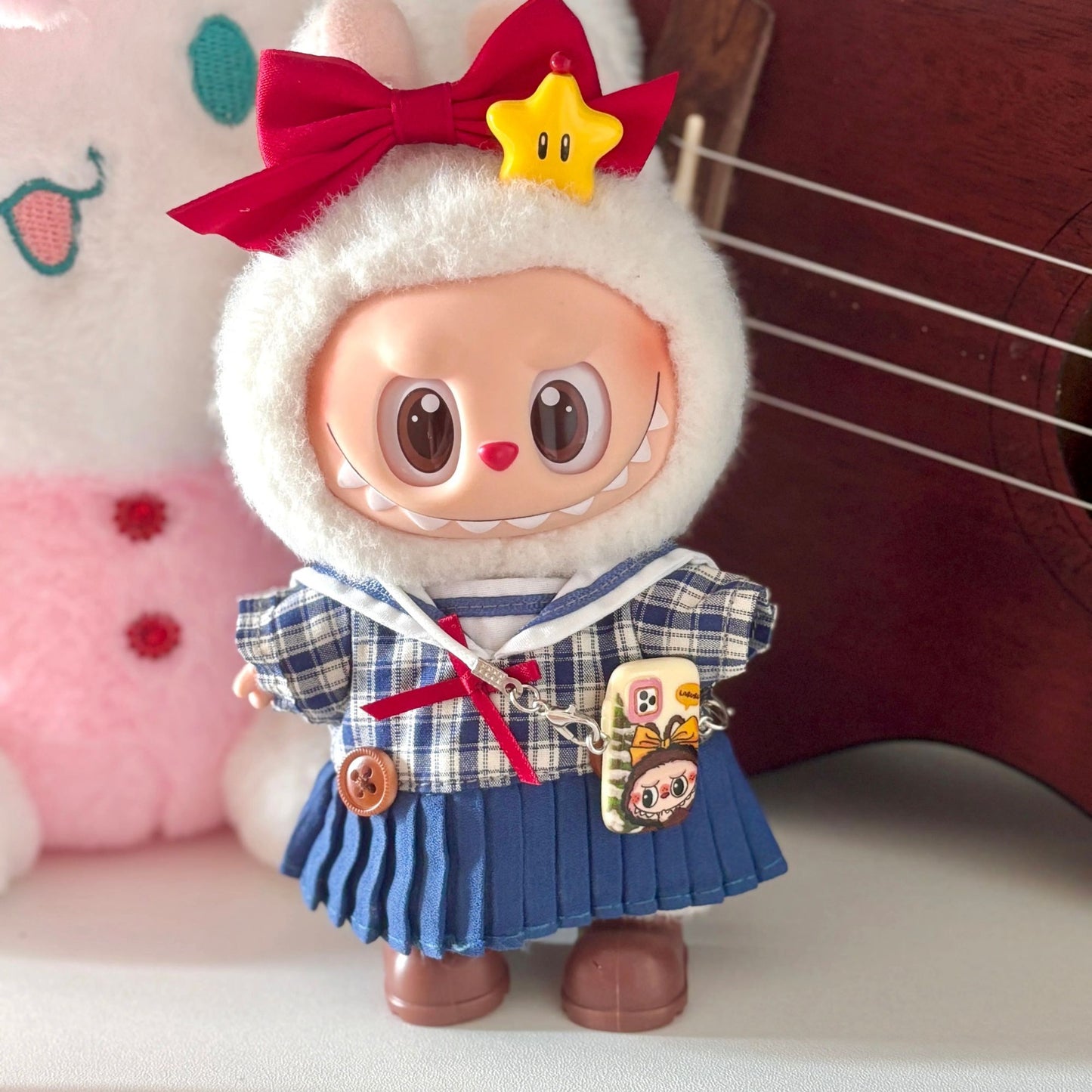 Labubu doll wearing handmade Japanese school uniform costume featuring navy and white plaid sailor-style top with white collar, blue pleated skirt with suspender straps, red bow tie and matching hair bow, yellow star hair clip, and character charm accessories. Perfect academic-themed doll outfit for kawaii fashion collectors and Japanese culture enthusiasts.