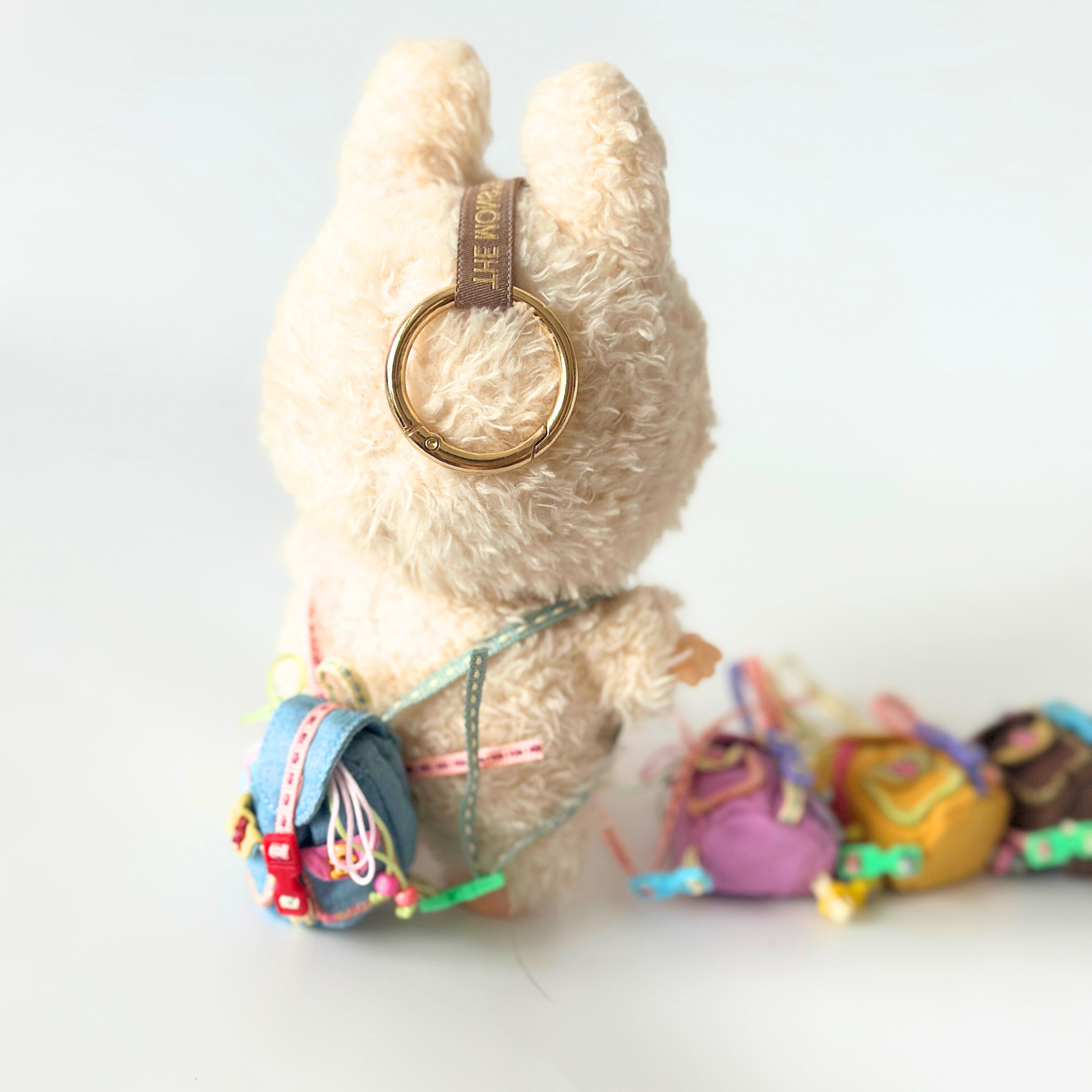 Labubu doll wearing blue corduroy miniature backpack with rainbow straps and metal buckle details, gold ring accessory on head, set against blurred background of colorful backpack collection in various colors including purple, yellow, and brown. Lifestyle photography showcasing handmade doll accessories and miniature backpack collection for collectors and toy enthusiasts