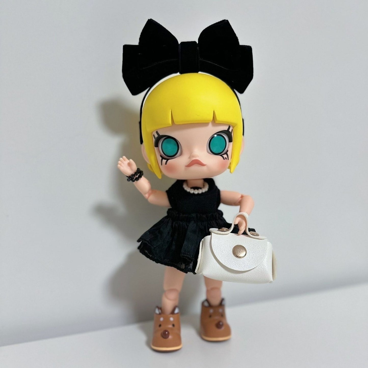 Molly collectible figure in a black ruffled dress with pearl necklace, black bow headband, waving with one hand while holding a white handbag in the other. The doll has blonde hair with bangs, large teal eyes, and brown boots, posed against a white background