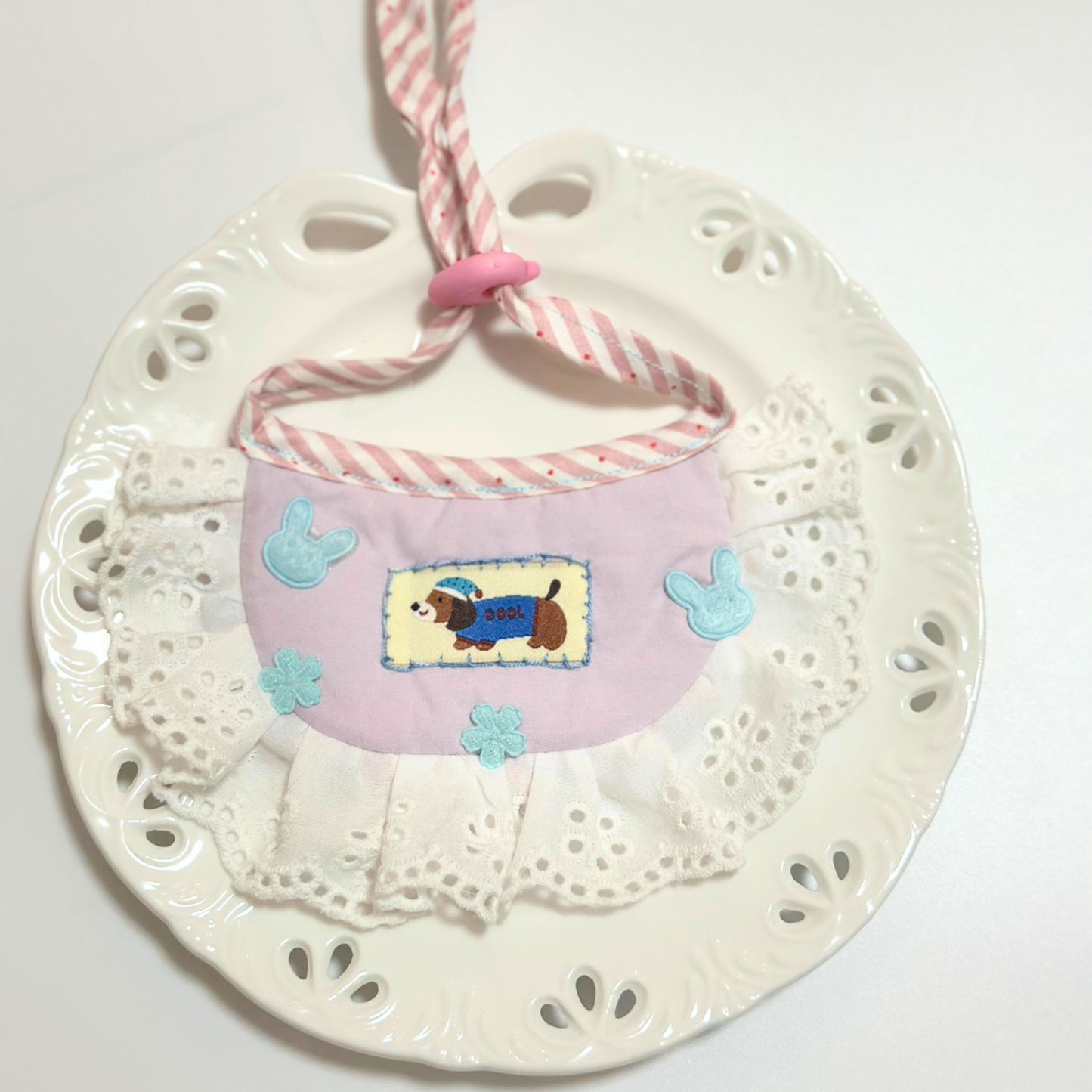 Dog bib with pom-pom trim on decorative plate - Light blue pet bandana bib with bunny pattern, ribbon bow, eyelet lace ruffle, styled on white paw print plate