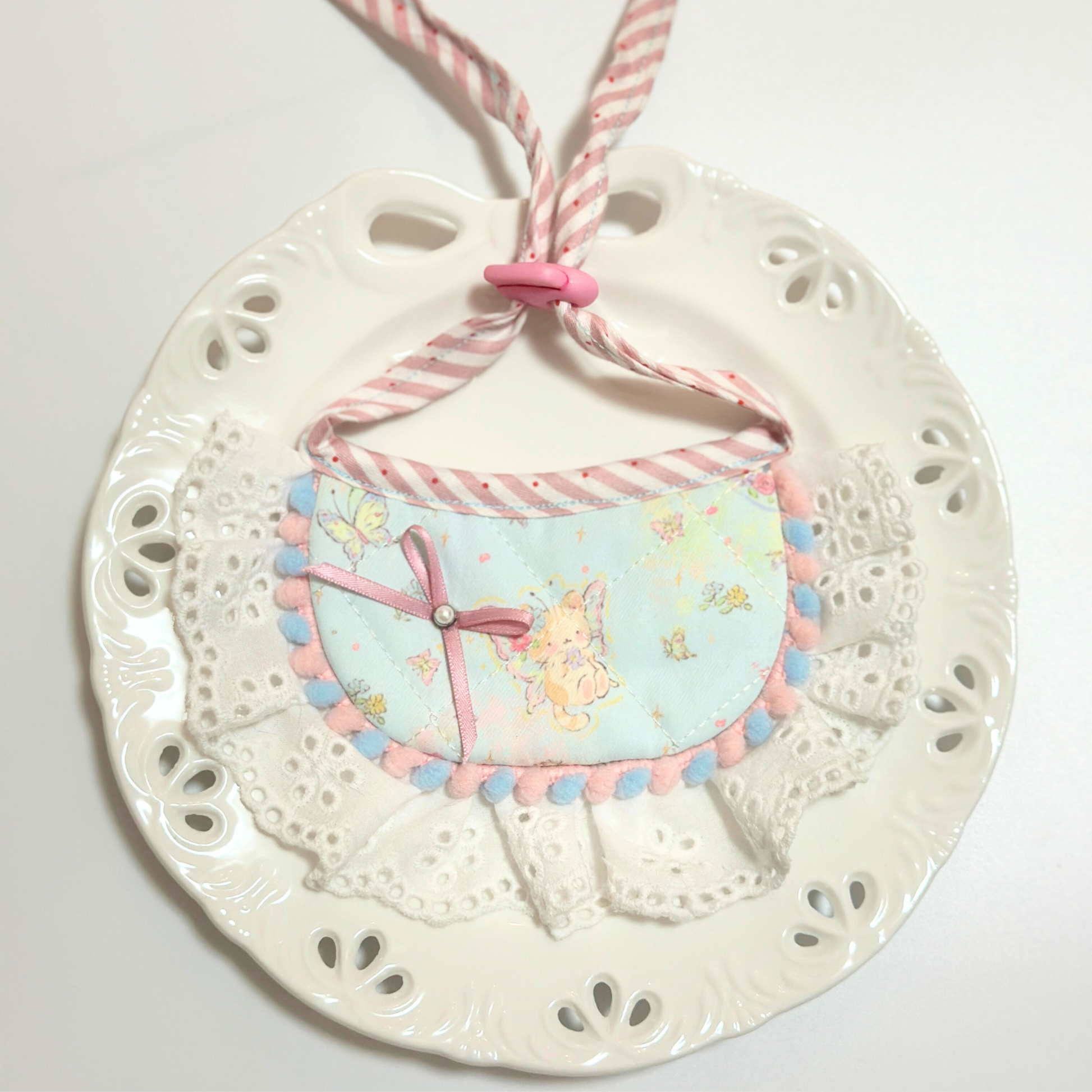 Dog bib with pom-pom trim on decorative plate - Light blue pet bandana bib with bunny pattern, ribbon bow, eyelet lace ruffle, styled on white paw print plate