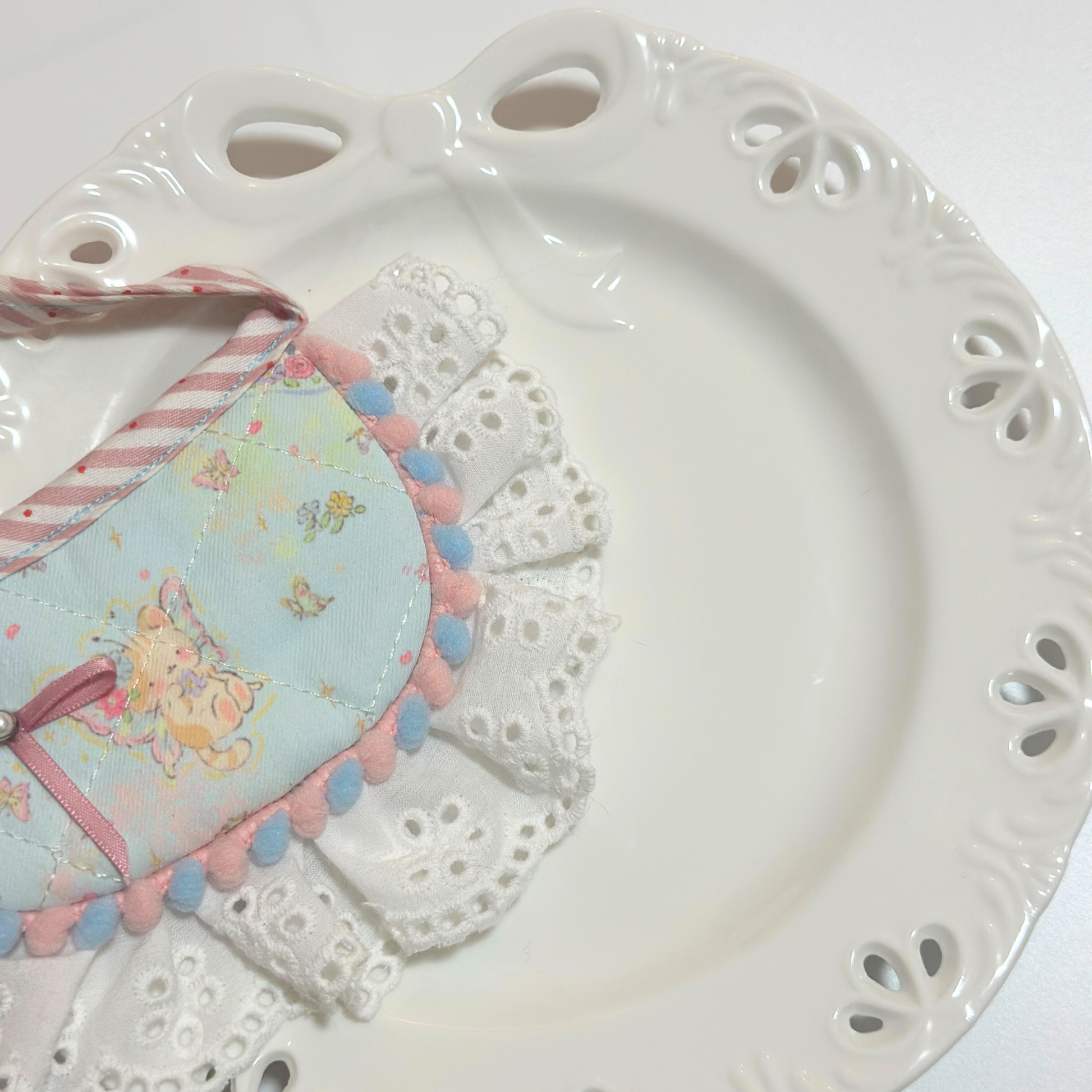 Dog bib with pom-pom trim on plate - Light blue pet bandana bib with bunny pattern, ribbon bow decoration, eyelet lace ruffle, displayed folded on white paw print plate