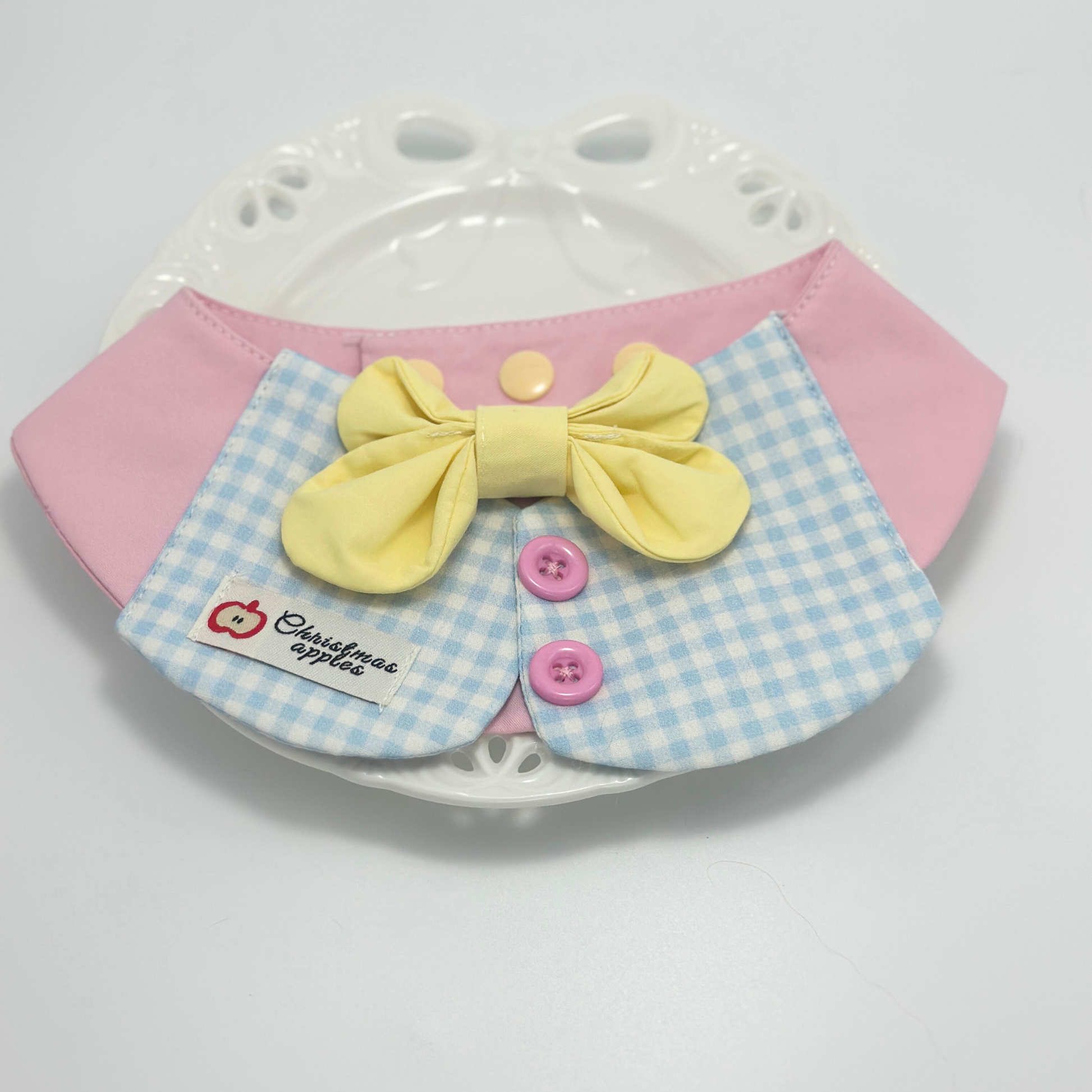 Pink pet bib with blue gingham collar and yellow bow tie displayed on white decorative plate with paw print pattern, featuring "Christmas apples" label and pink buttons - elegant pet accessory presentation