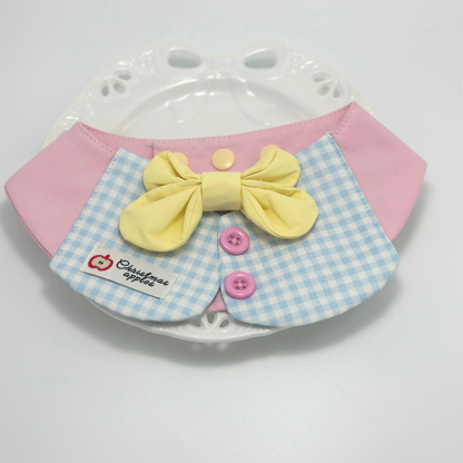 Pink pet bib with blue gingham collar and yellow bow tie displayed on white decorative plate with paw print pattern, featuring "Christmas apples" label and pink buttons - elegant pet accessory presentation