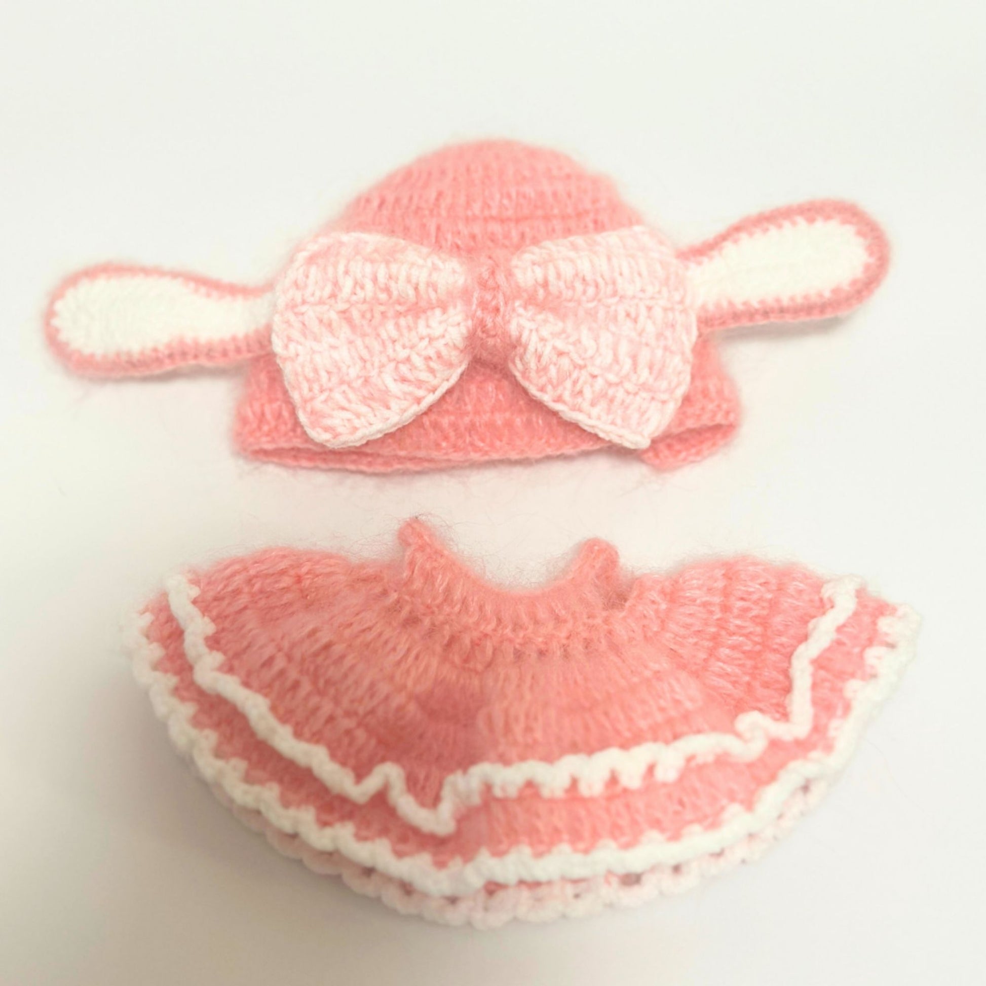 Handmade knitted bunny costume set for Labubu doll laid flat on white background. Top piece shows pink rabbit ears beanie with white bow decoration and long ear flaps. Bottom piece displays matching pink ruffled dress with white scalloped trim and layered design. Complete two-piece coral pink knitted outfit with white accent details.