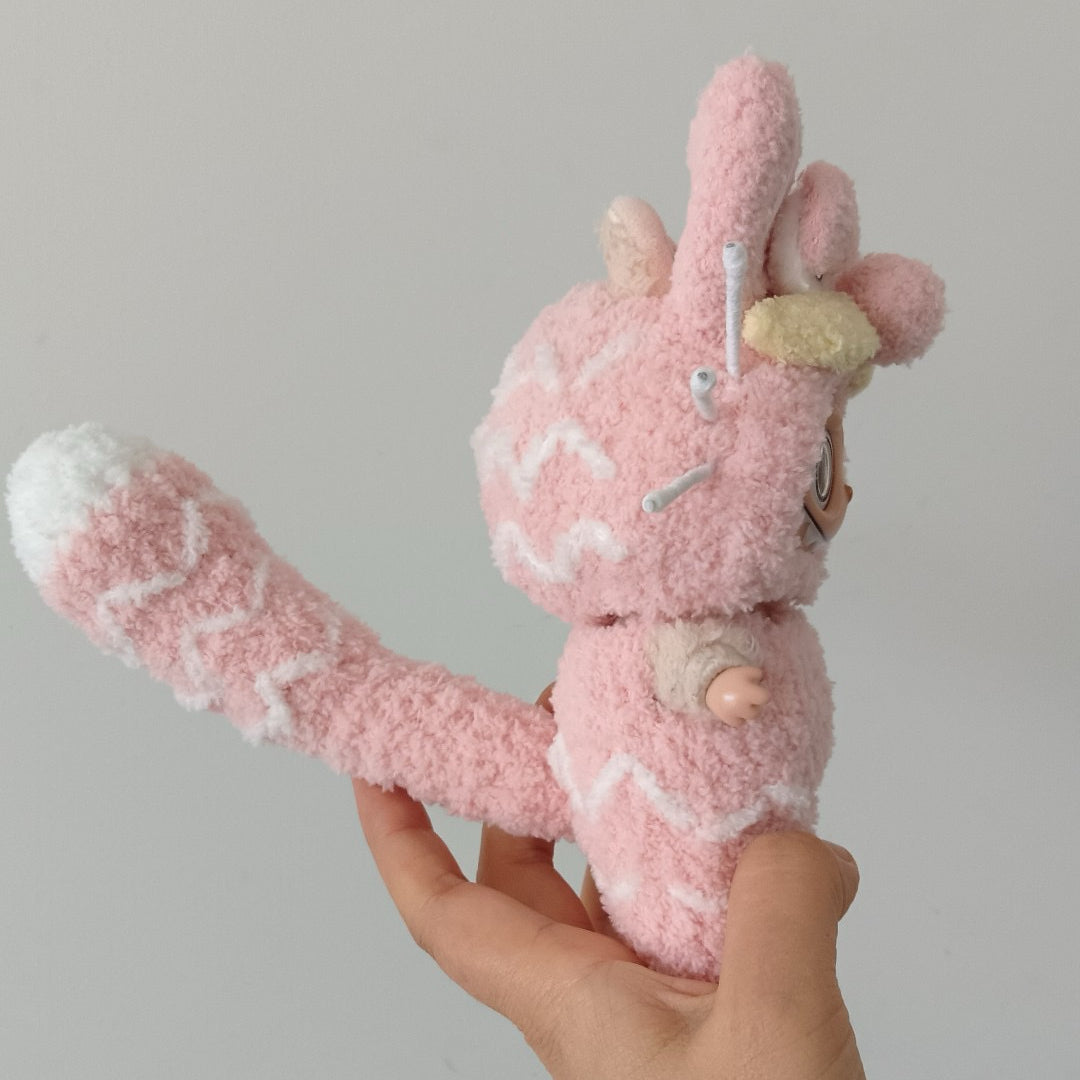 sideview of labubu wearing a pink garfield style outfit with white patterns held by a hand against a gray background