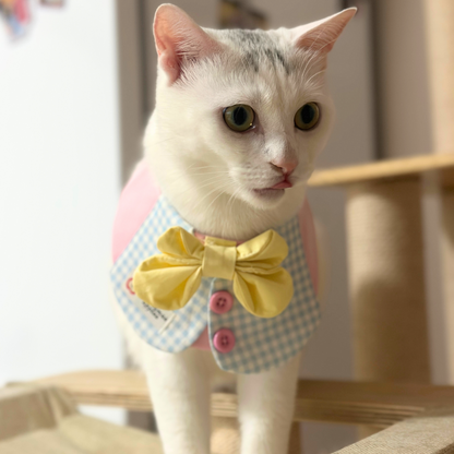 White cat with gray markings and green eyes wearing pink and blue gingham pet bib with yellow bow tie, sitting indoors and looking directly at camera - adorable pet portrait with formal accessories