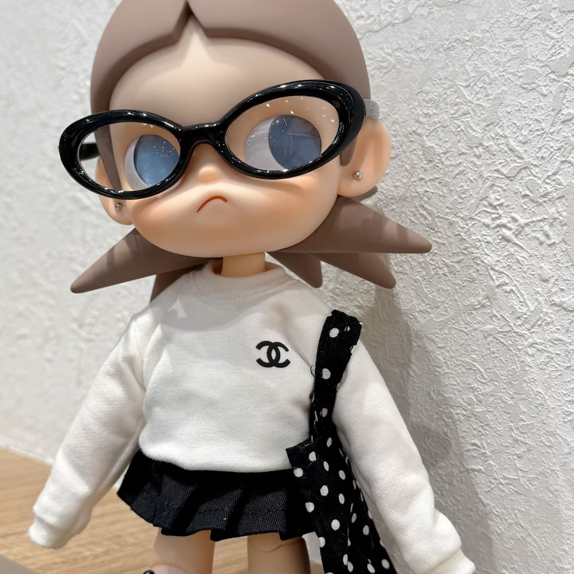 Zsiga doll wearing Chanel-style white sweater with black logo, black pleated skirt, polka dot bagsand oversized black sunglasses