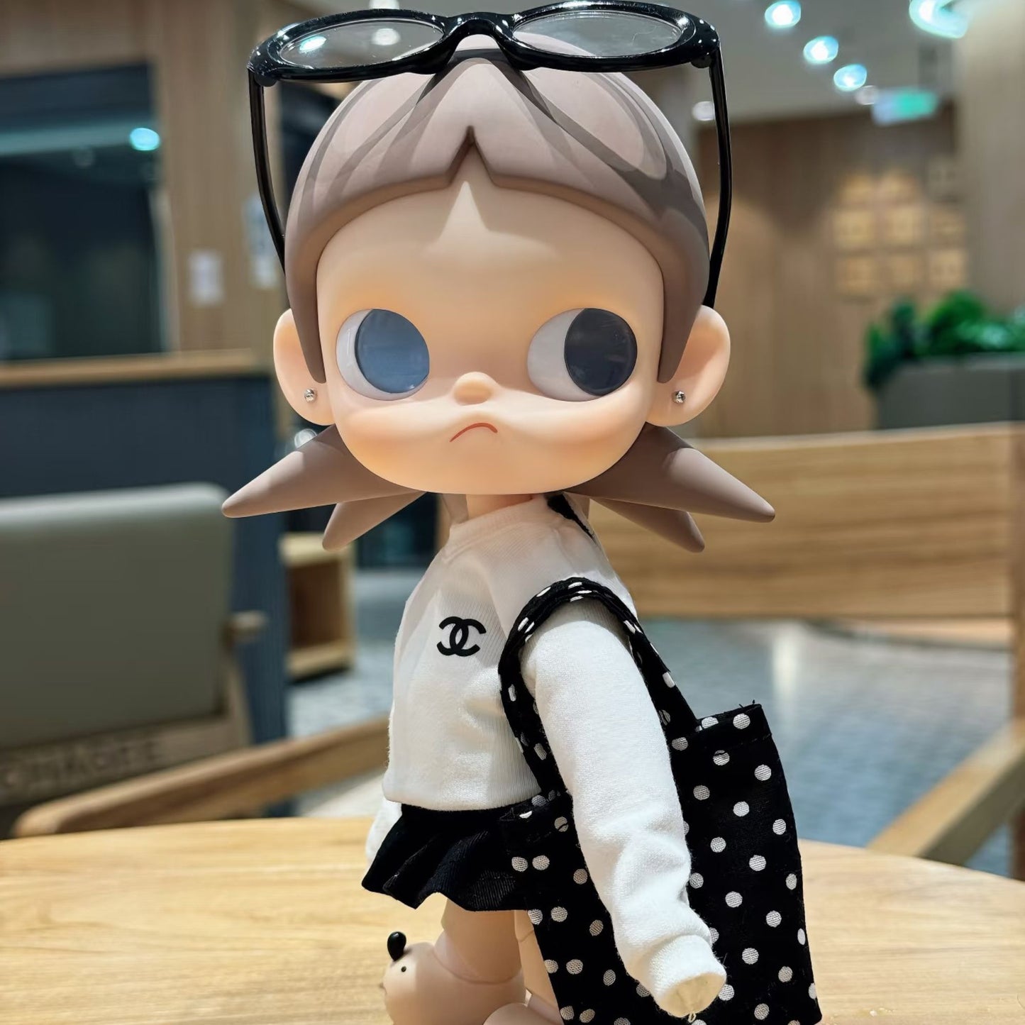 Zsiga doll in Chanel-style white sweater and black pleated skirt with polka dot bag and black sunglasses on head - lifestyle photo in modern cafe setting